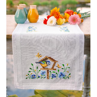 Tischläufer mit buntem Vogelhäuschen und Blumenarrangement auf einem Tisch.