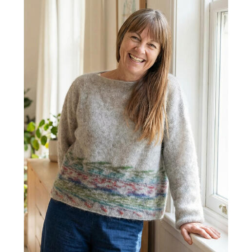 Frau lächelt in einem grauen Pullover mit buntem Muster, steht vor einem Fenster.