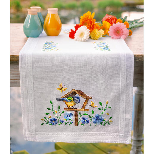 Tischläufer mit buntem Vogelhäuschen und Blumenarrangement auf einem Tisch.