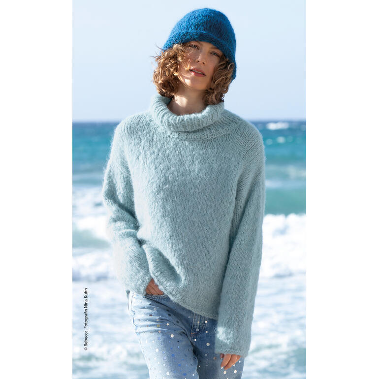 Frau in einem hellblauen Pullover mit hohem Kragen und Mütze, steht am Strand.