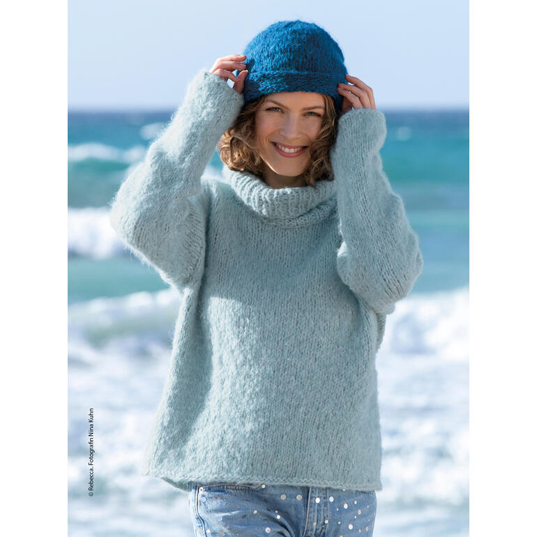 Frau in einem hellblauen Pullover mit hohem Kragen, trägt eine Mütze am Strand und lächelt.