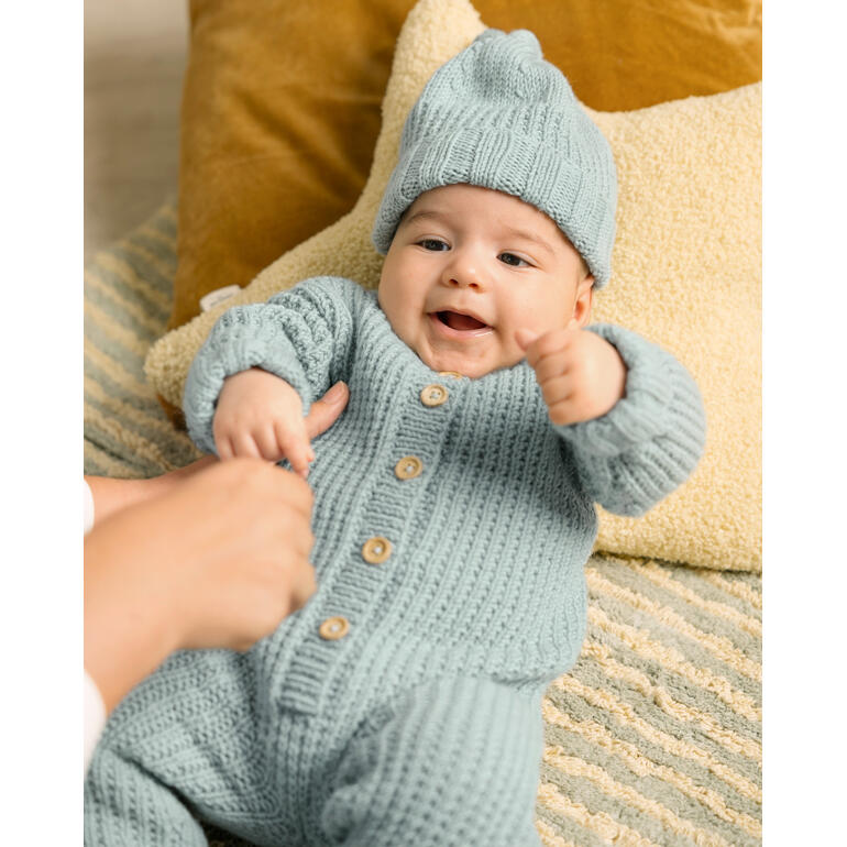 Baby in hellblauem Strampler und Mütze, spielt auf einem Teppich.