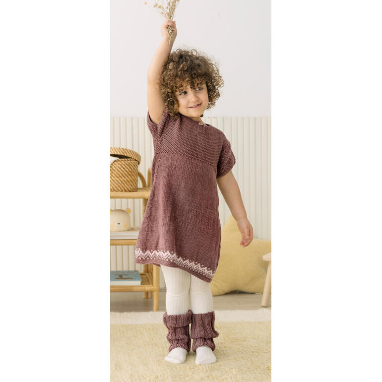 Kleines Kind in lila Kleid mit Stulpen, lächelt mit einer Blume in der Hand.
