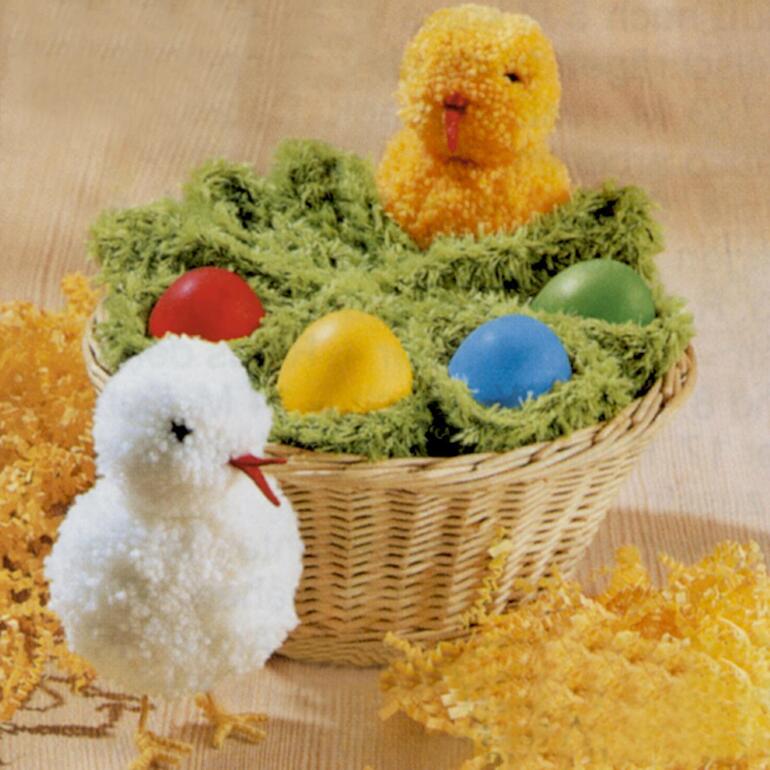 Zwei Pompon-Küken in einem Korb mit bunten Eiern auf grünem Gras.