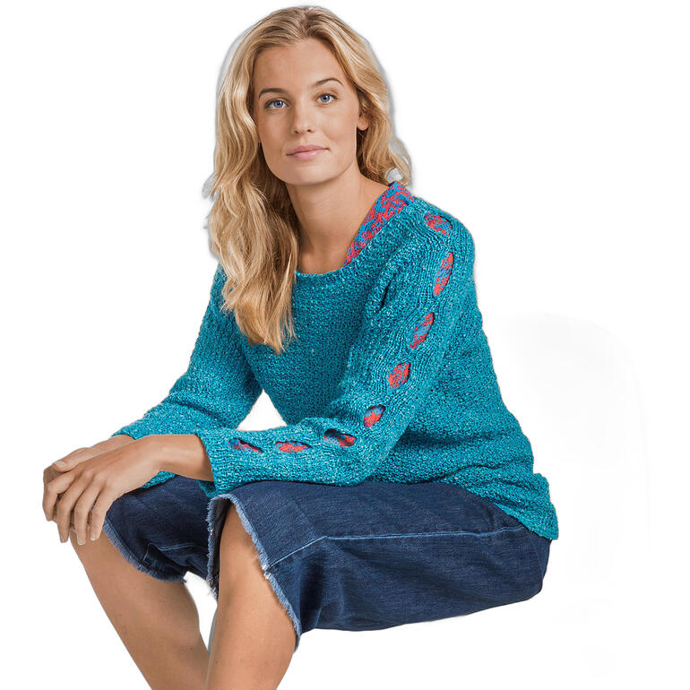 Frau in türkisfarbenem Pullover mit dekorativen Details auf einer Sitzgelegenheit, schaut in die Kamera.