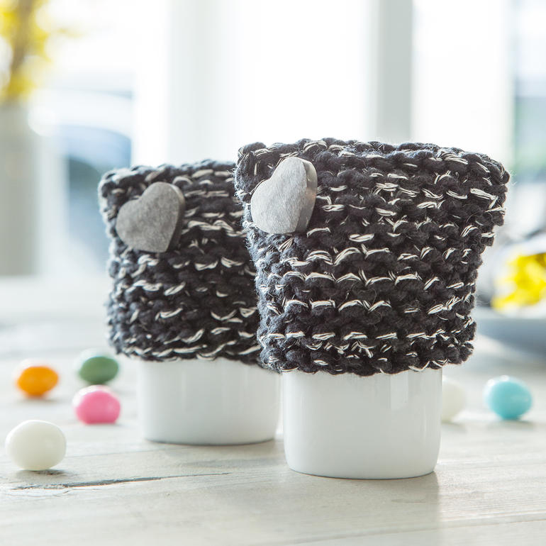 Zwei Eierwärmer aus dunkelgrauer Wolle mit Herzen, auf einem Tisch.