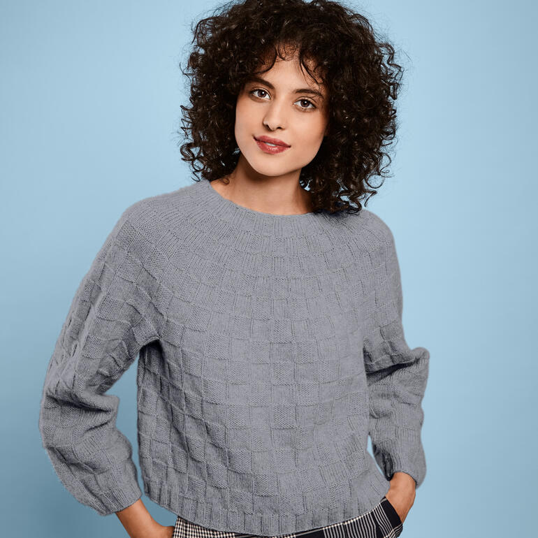 Frau mit lockigen, dunklen Haaren trägt einen grauen Pullover mit strukturierter Oberfläche.