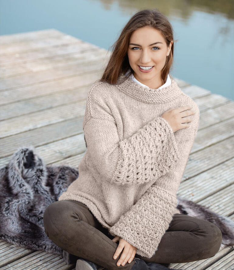 Frau in einem beigen Pullover sitzt auf einem Holzsteg am Wasser, mit lächelndem Gesicht.