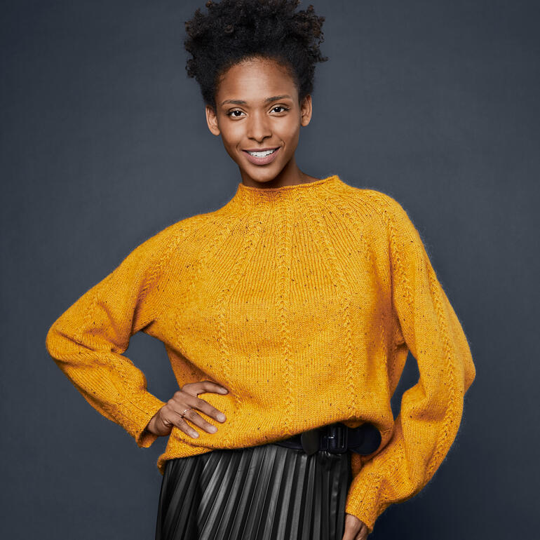 Frau trägt einen ockergelben Pullover aus Como Tweed, posiert lächelnd in freundlicher Haltung.