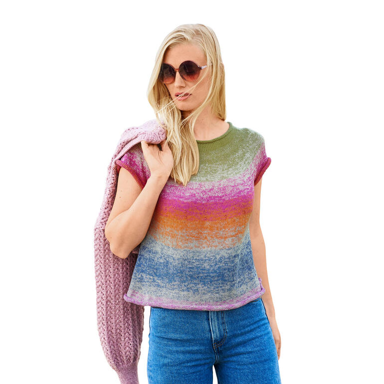 Frau in einem bunten Top mit kurzen Ärmeln und Jeans, lächelt vor hellem Hintergrund.