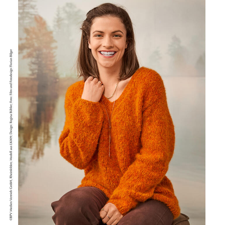Lächelnde Frau in orange Pullover mit V-Ausschnitt vor einer Waldlandschaft.