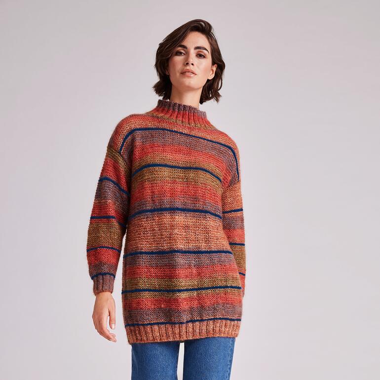 Frau in buntem Pullover mit gestreiftem Muster steht vor neutralem Hintergrund.