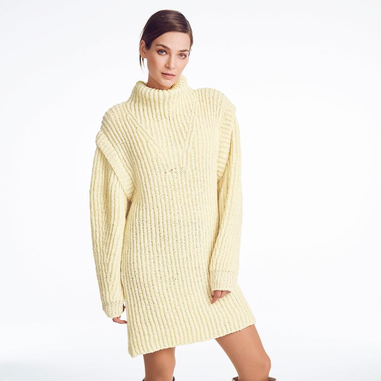 Frau in oversized, beigefarbenem Pullover mit hohem Kragen vor heller Wand