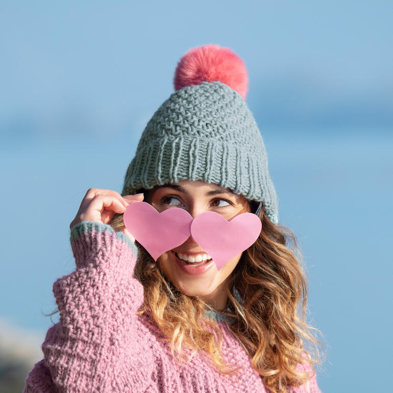 Frau trägt eine graue Mütze mit rosa Pompon und hält Herzbrille vor ihrem Gesicht.