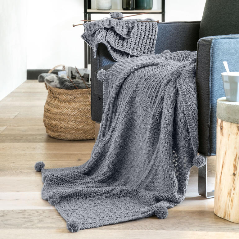 Grau gestrickte Decke mit Quasten, drapiert über ein Sofa mit einem Korb daneben.