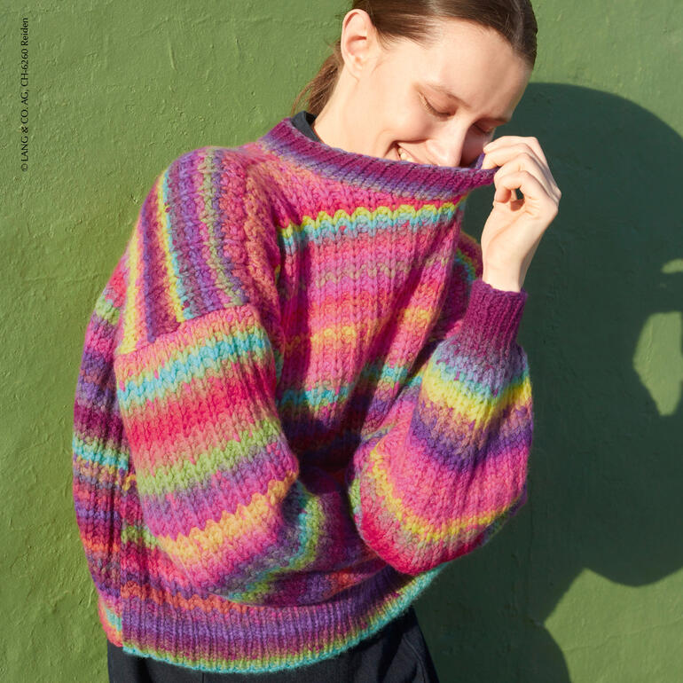 Frau mit einem bunten Pullover aus Mystery, lächelt und zieht den Kragen über ihr Gesicht.
