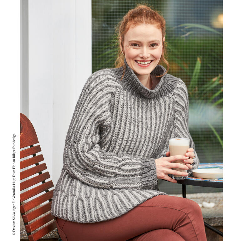 Lächelnde Frau trägt grauen Pullover mit Rollkragen und hält einen Becher in der Hand.