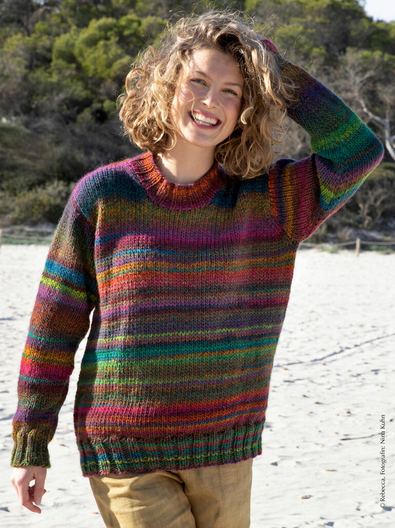 Frau trägt einen bunten, gestreiften Pullover am Strand und lächelt fröhlich.