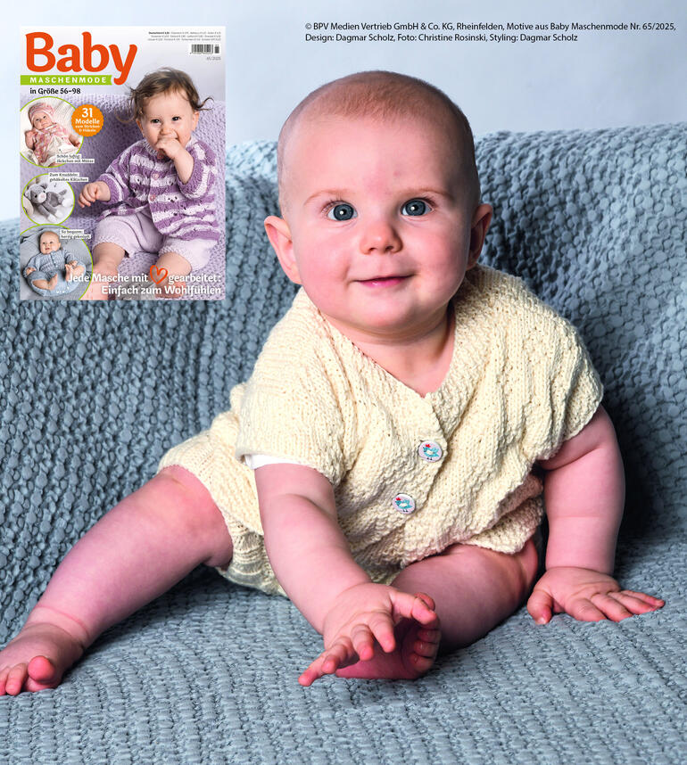 Baby mit hellen Strickwaren auf einem grauen Kissen, lächelt in die Kamera.