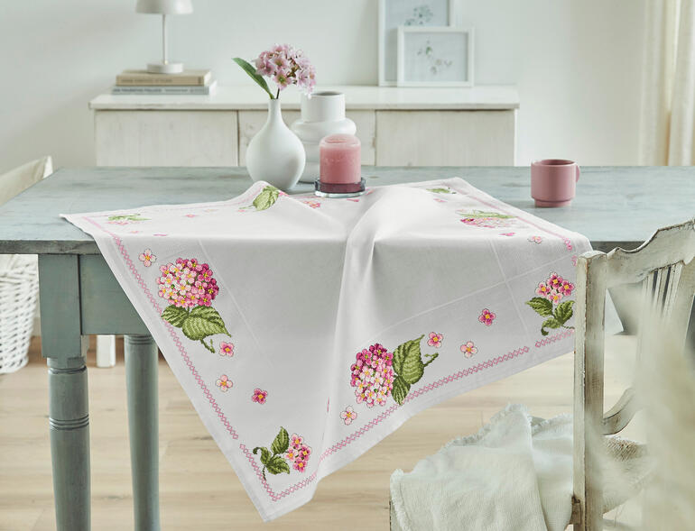 Aida-Tischdecke mit rosa Hortensien und zarten Blüten auf einem Tisch.