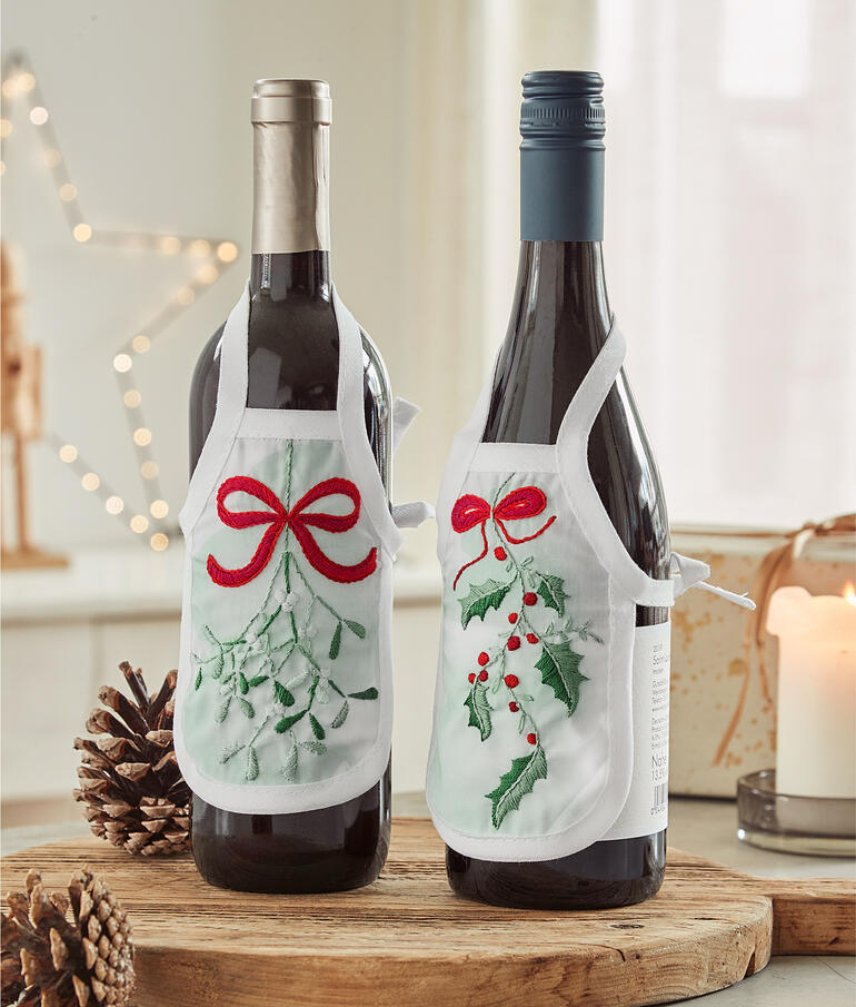 Zwei Flaschenschürzen mit Mistelzweig-Design an Weinflaschen.