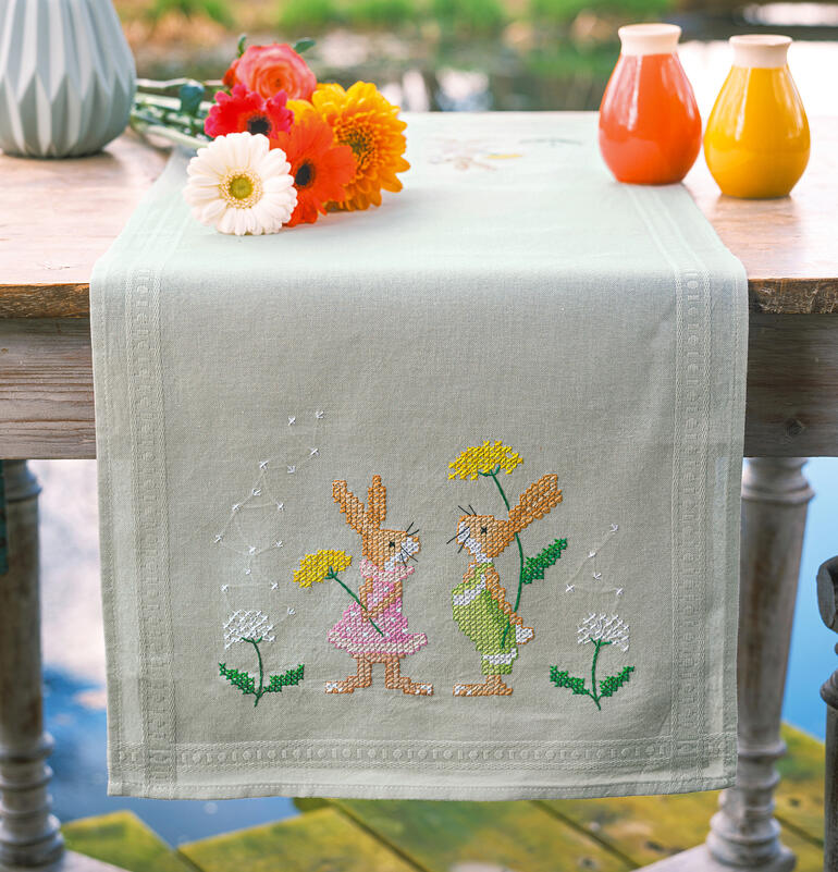 Tischläufer mit zwei Osterhasen, Blumen und bunten Vasen auf einem Tisch.